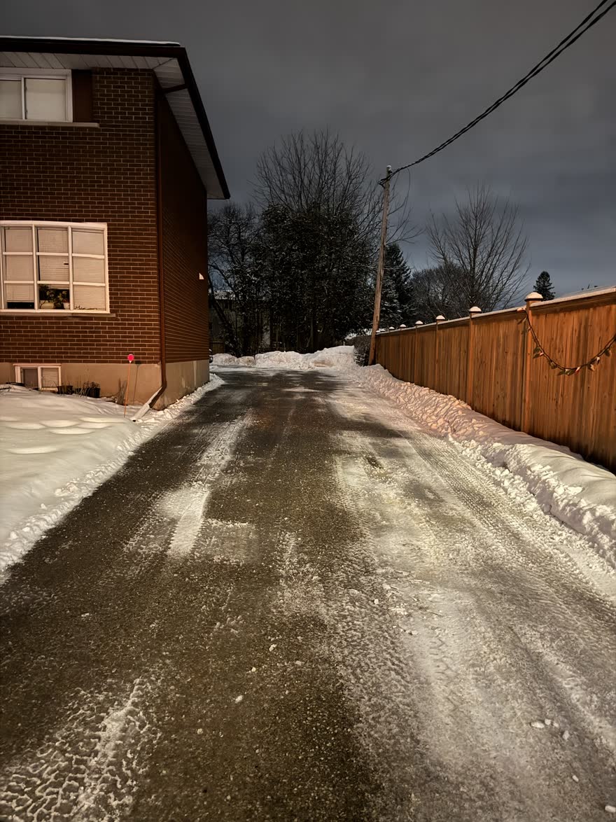 Long laneway beside a brick building at night, plowed edge to edge and salted, with a fence on one side